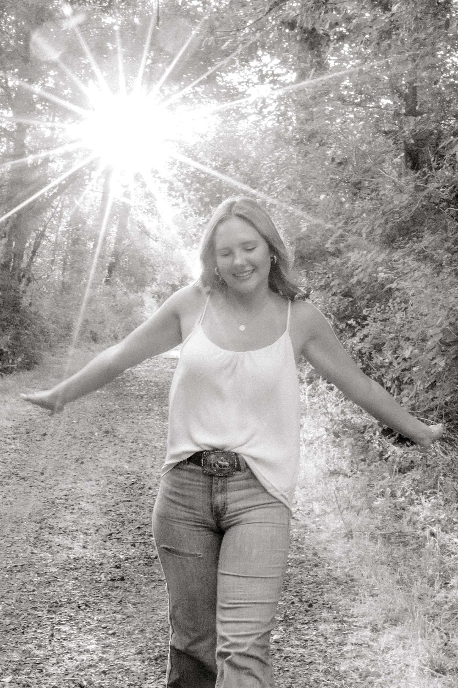 Black and white photo of a smiling woman with arms outstretched on a sunlit forest path wearing jeans and a tank top.