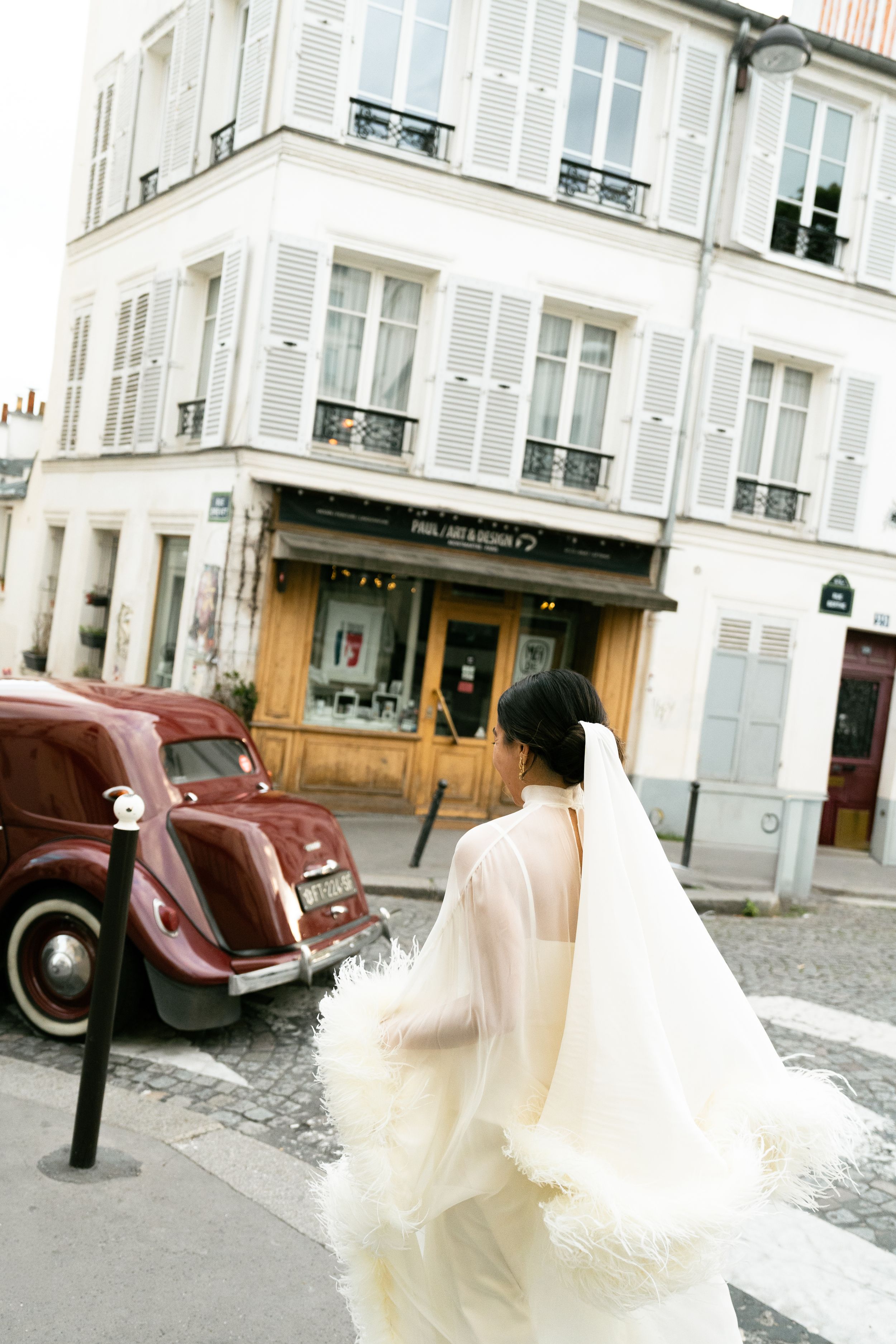Bride in flowing white gown and veil walks through charming Parisian street corner with vintage red car and classic buildings.
