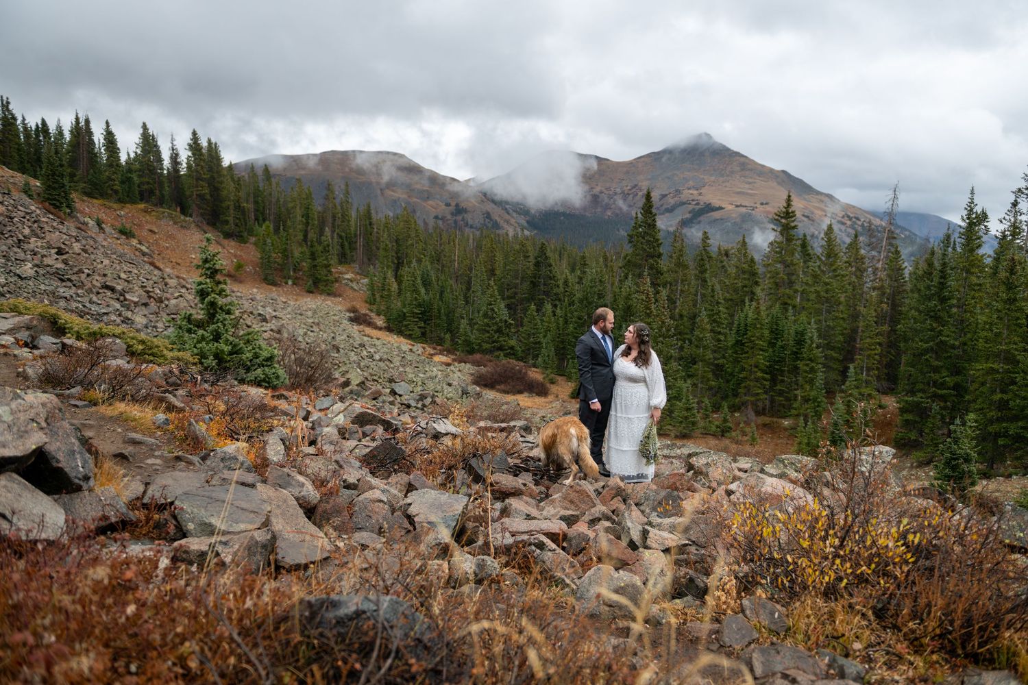 An eloping couple stand near each other with their dog at their feet in a dramatic mountain area with misty mountain peaks behind them.