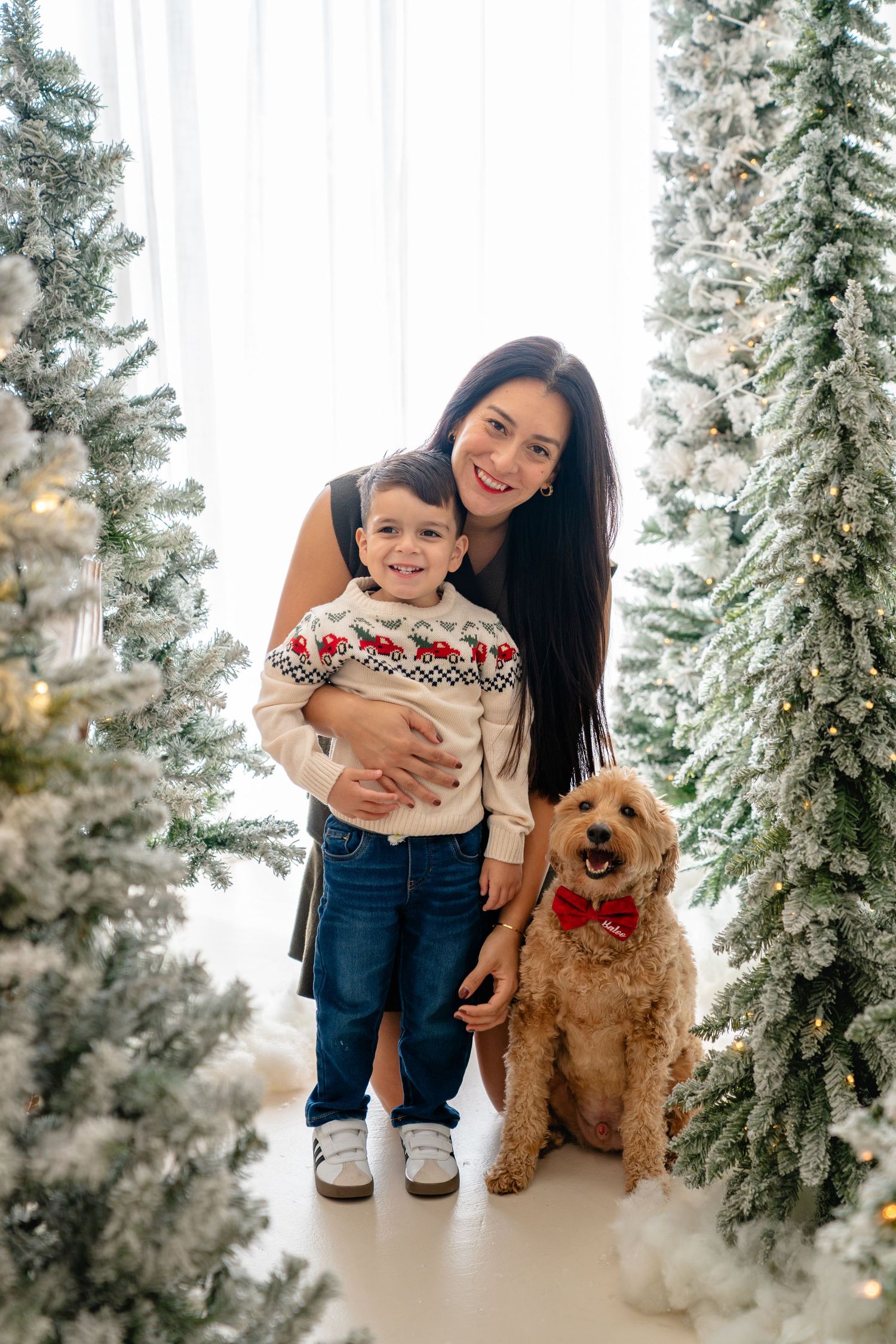 Woman and young boy in Christmas sweater pose with golden doodle dog among snow-flocked Christmas trees.