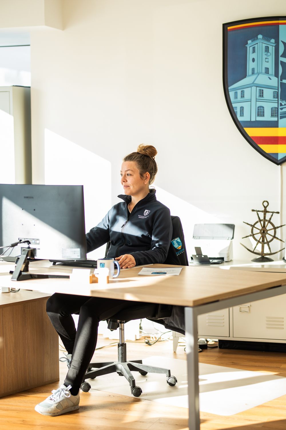 Woman in navy jacket working at a computer desk in a sunlit office with a shield logo on the wall.