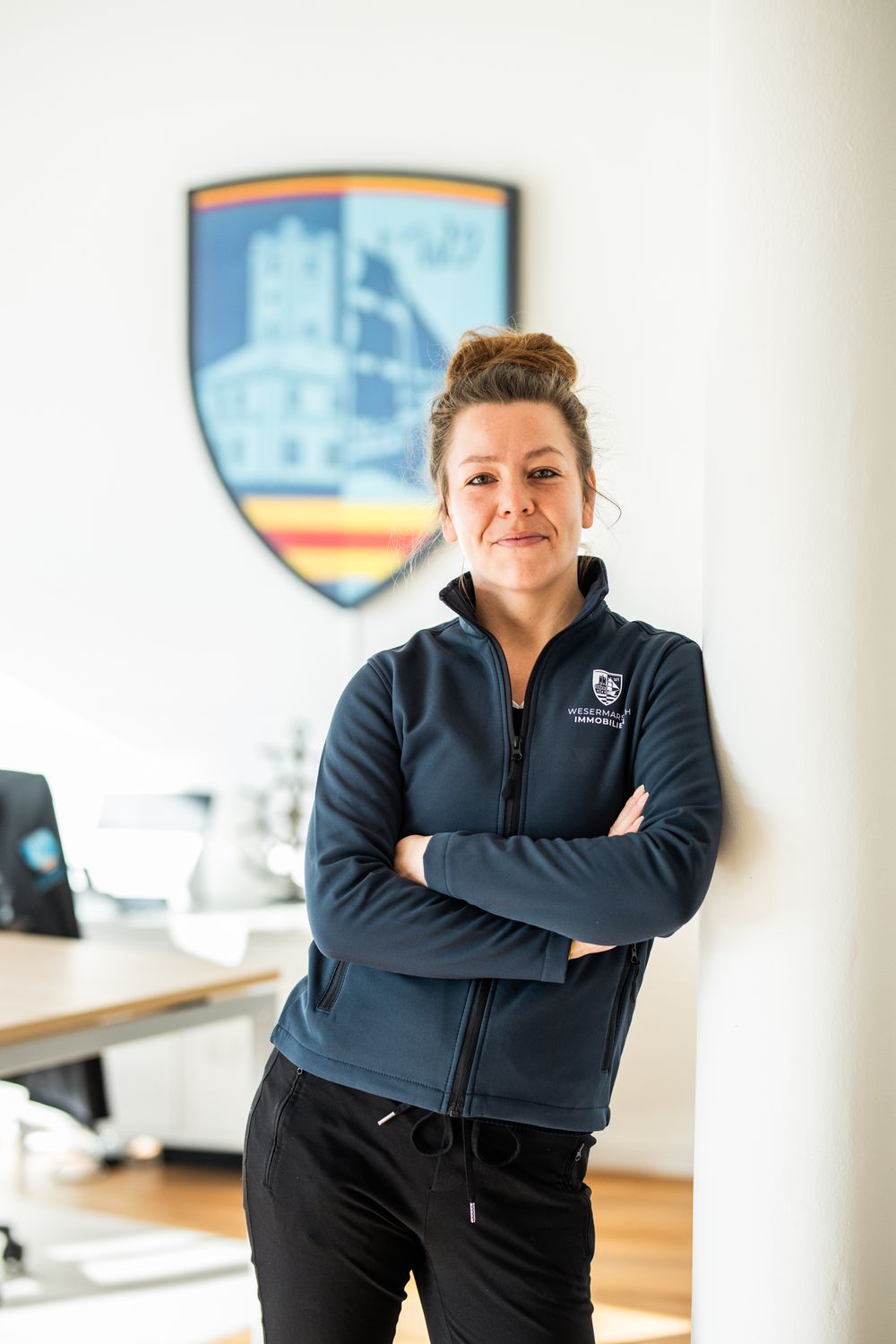 Woman in Wesermarsch Immobilien fleece jacket standing with arms crossed in front of a branded shield logo.