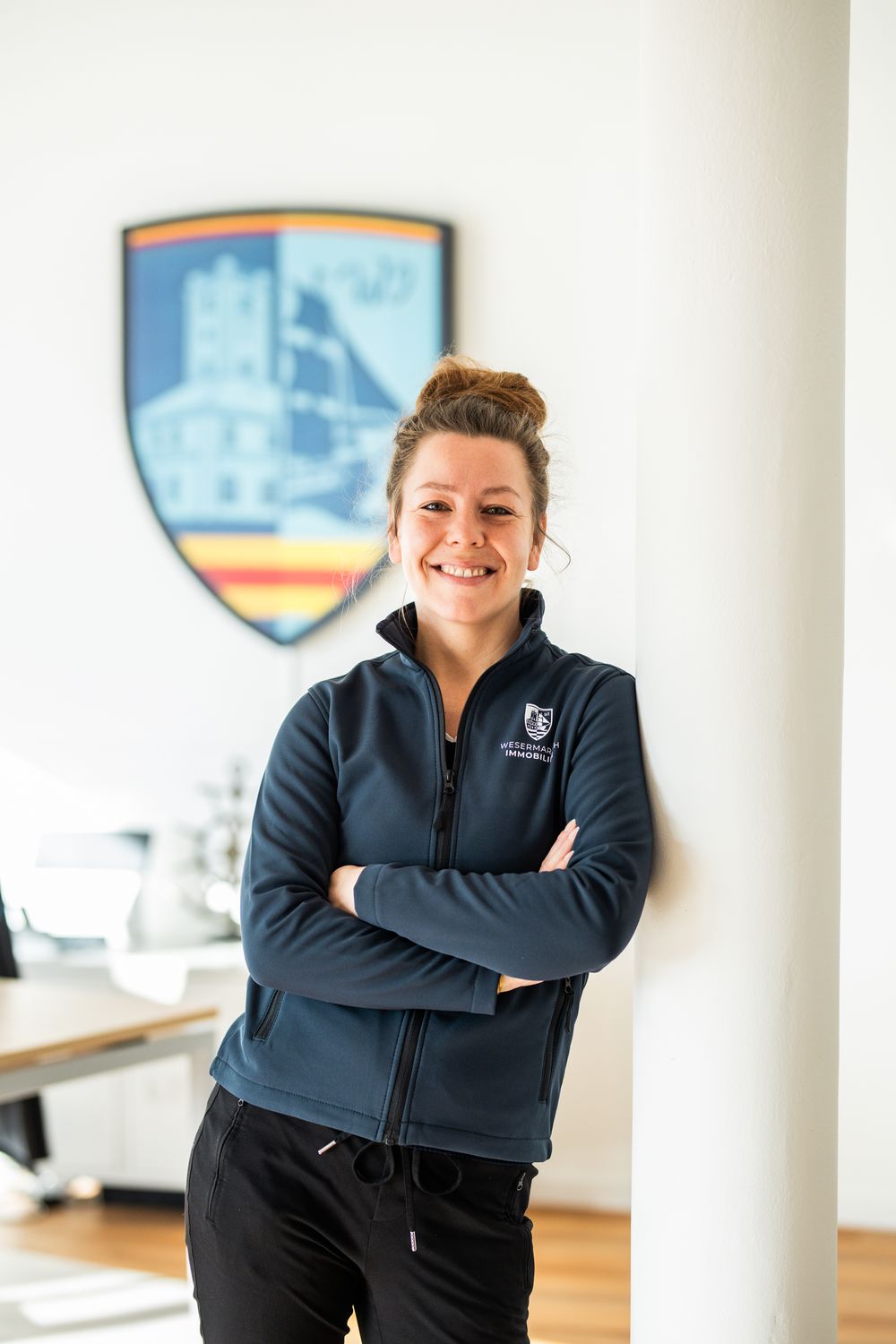 Smiling woman in navy Wesermarsch Immobilien fleece jacket standing with arms crossed in a bright office.