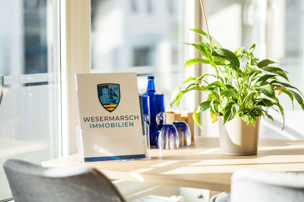 Wesermarsch Immobilien branded sign on a sunny windowsill beside a green plant and blue decorative bottles.