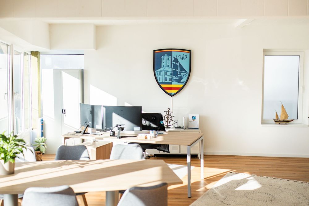 Sunlit Wesermarsch Immobilien office interior with wooden desks, gray chairs, and a large shield logo on the wall.