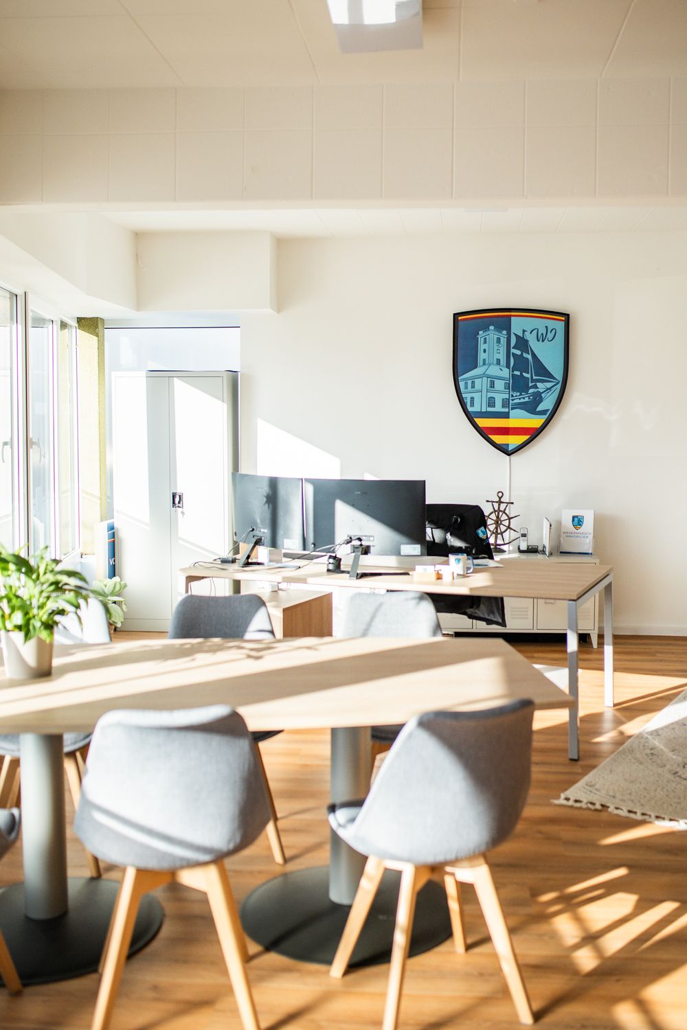 Bright modern real estate office with gray chairs, wooden table, and Wesermarsch Immobilien shield logo on wall.