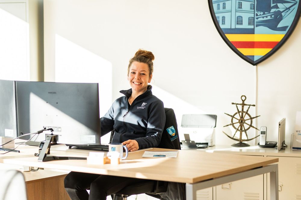 Smiling woman in uniform sitting at office desk with computer, anchor decor, and city coat of arms on wall.