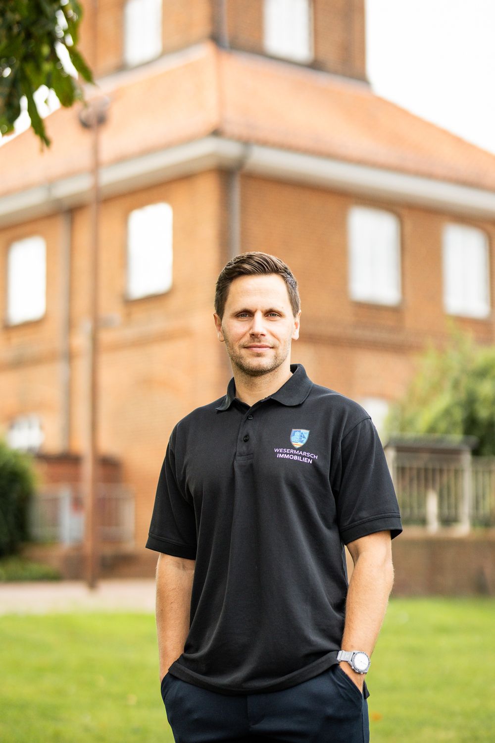 Man in black Wesermarsch Immobilien polo shirt standing outdoors in front of a red brick building.
