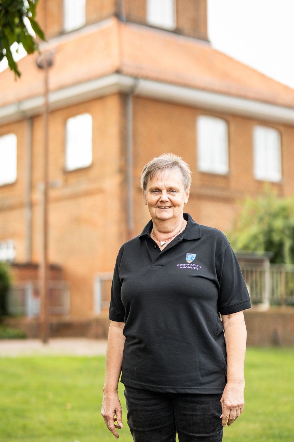 Smiling woman in black Wesermarsch Immobilien polo shirt standing on grass in front of a red brick building.