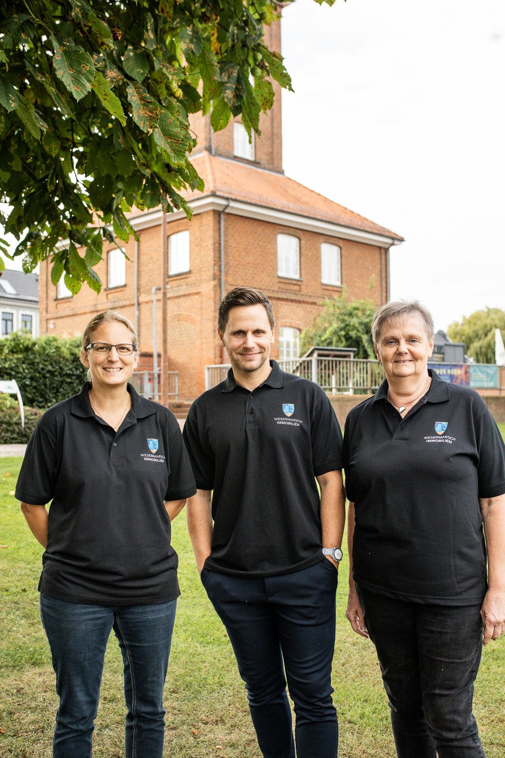 Three smiling team members in black polo shirts standing outdoors in front of a red brick building.