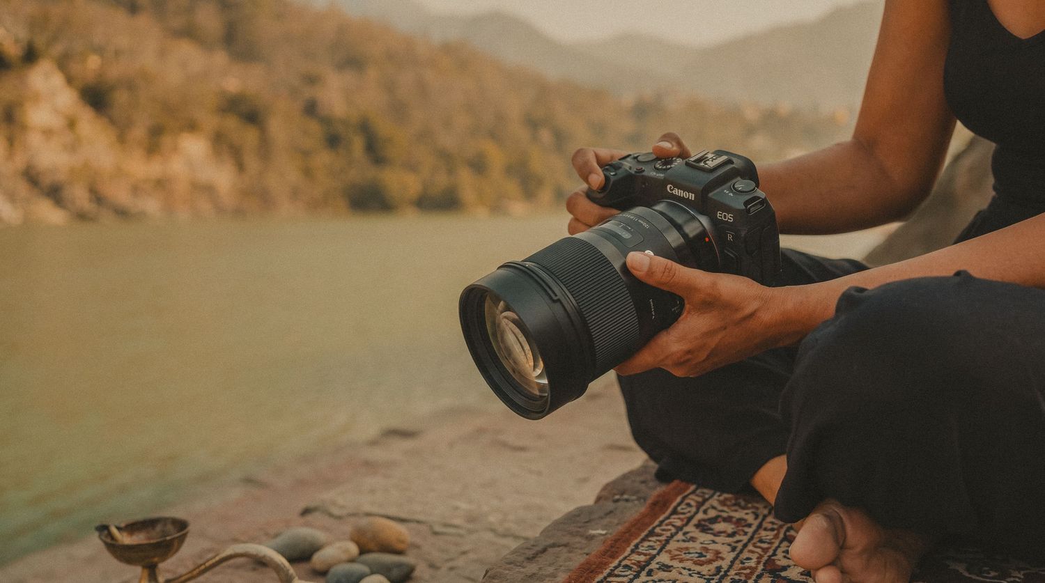 Photographer holds a professional DSLR camera with large lens while sitting outdoors near a mountain lake.