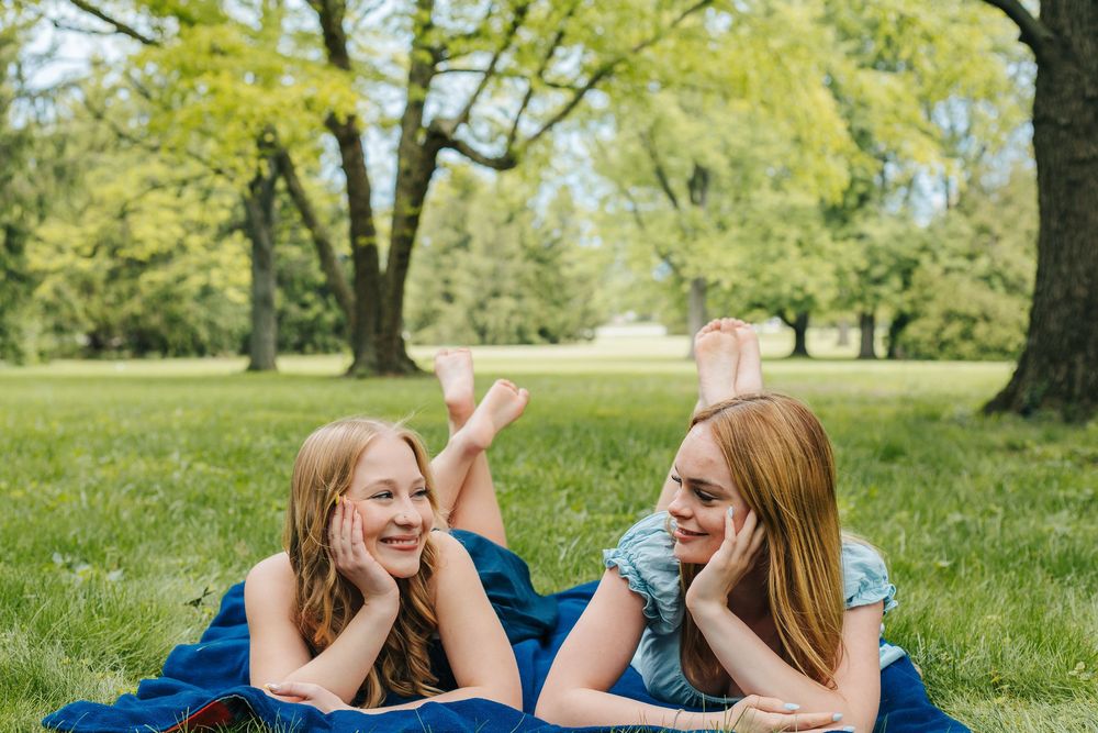 Two girls lying on a blue blanket in a park, smiling at each other surrounded by lush green grass and trees.