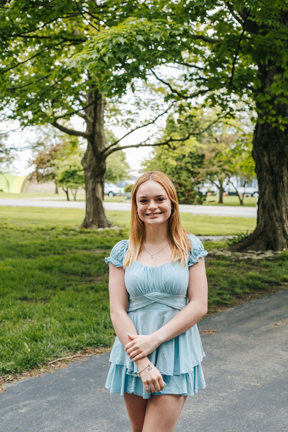 Young woman in light blue mini dress smiling in a green park with large trees.