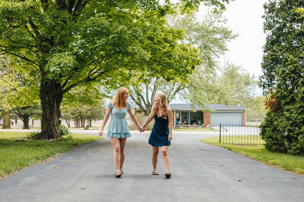 Two girls holding hands walking down a tree-lined driveway in summer dresses.