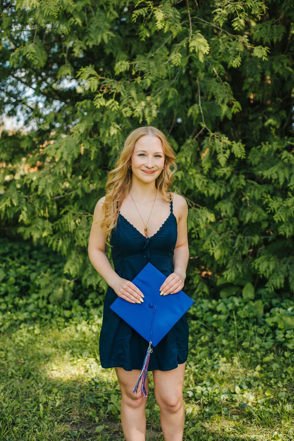 Smiling blonde graduate in navy dress holds blue graduation cap outdoors among lush green trees.