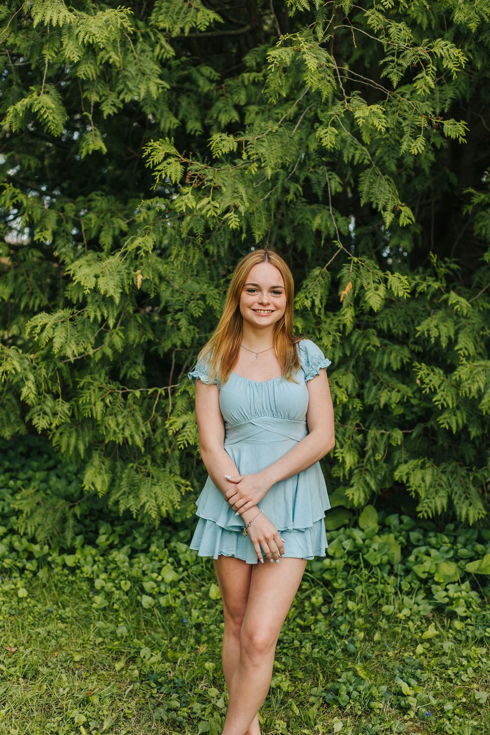Smiling young woman in teal ruffle mini dress standing before lush green foliage.