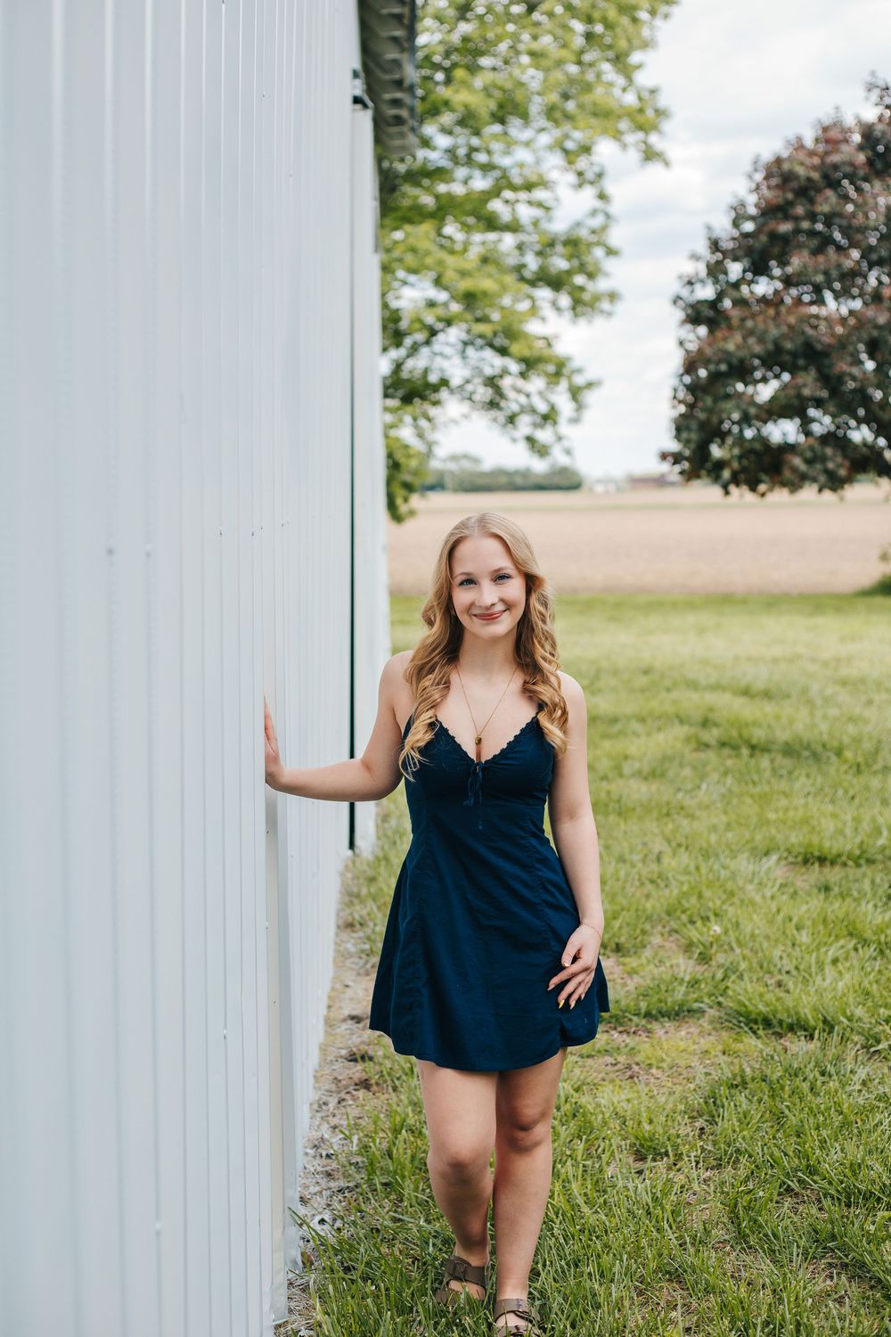 Young woman in navy blue dress posing by white barn wall in rural setting with green grass and trees.