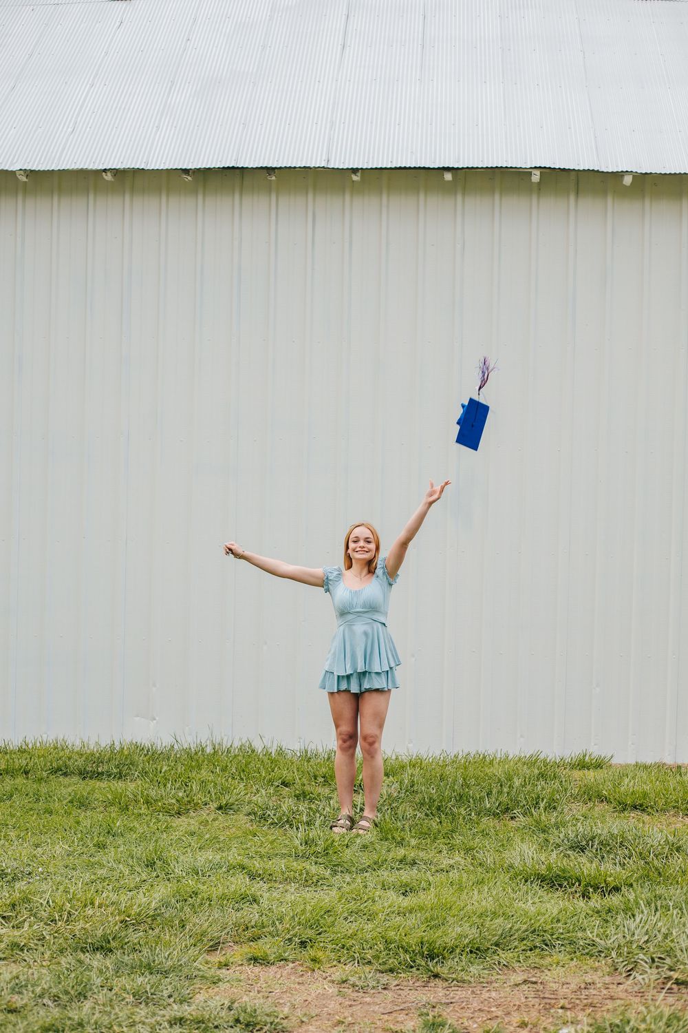Happy graduate in blue dress tossing graduation cap in the air outside a barn.