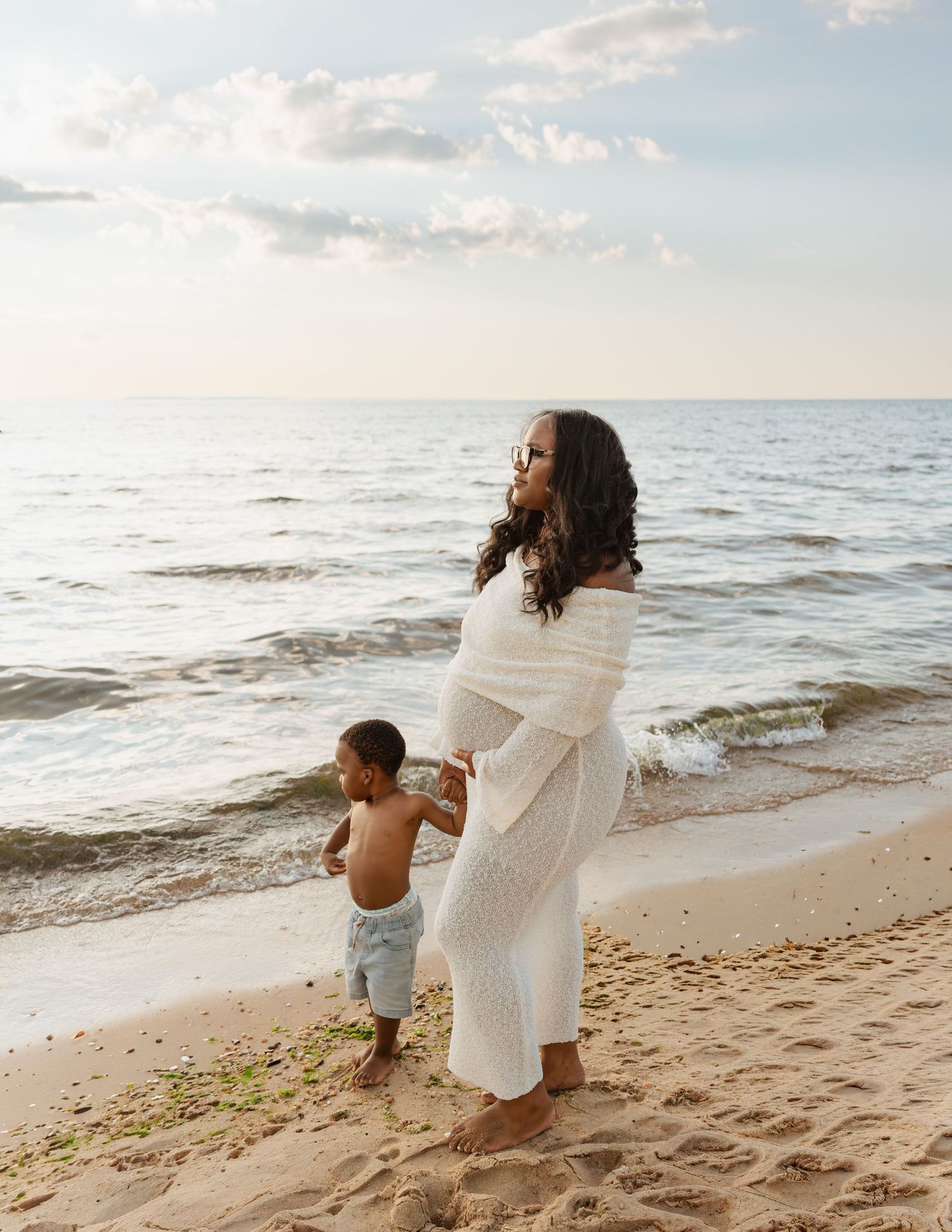Pregnant woman in white dress stands barefoot on sandy beach with toddler boy, ocean waves behind them.