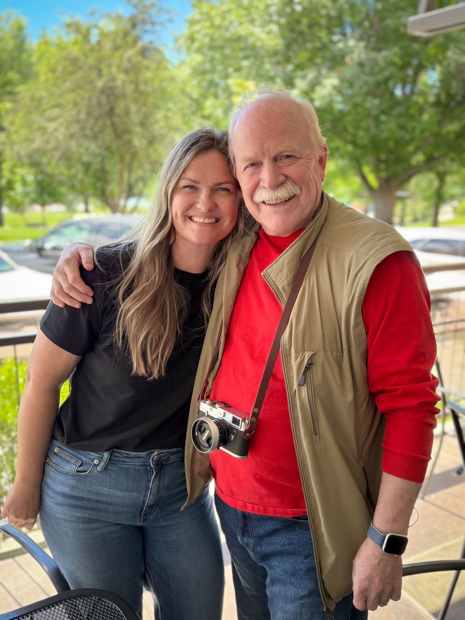 Two smiling people pose together outdoors, man wearing red shirt, tan vest, and camera, woman in black top and jeans.