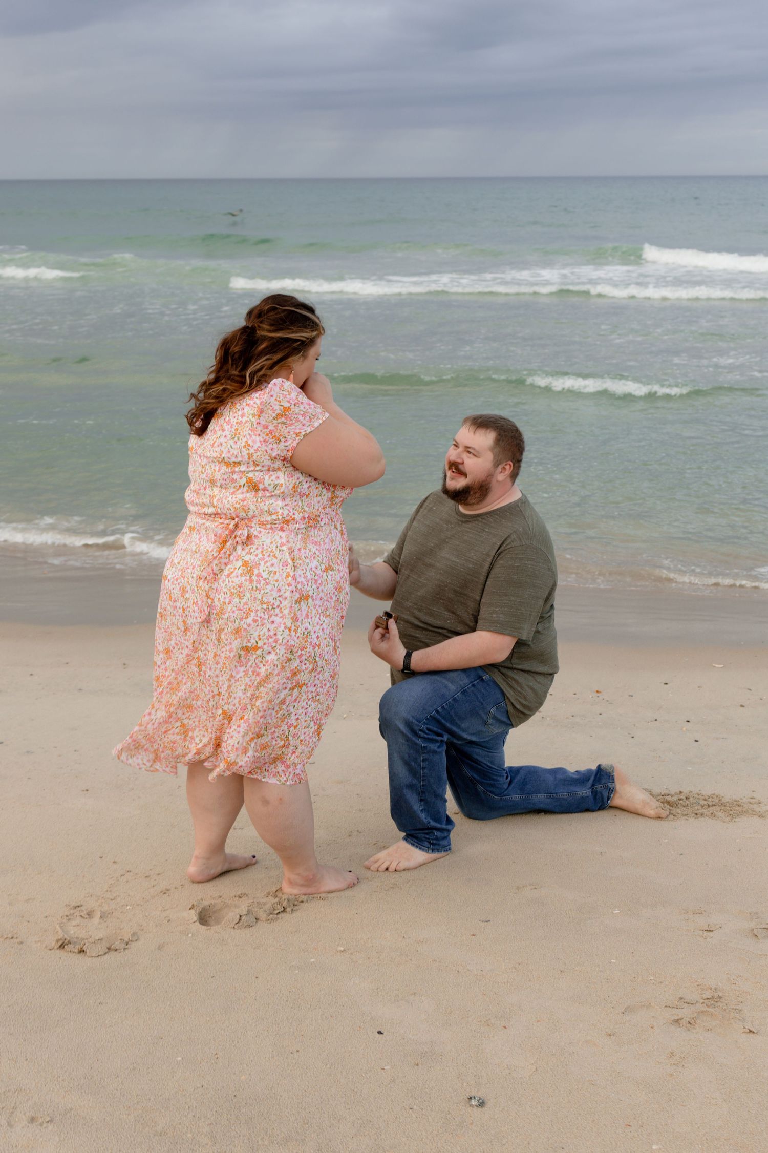 Man proposes on bended knee to woman in floral dress on sandy beach with ocean waves in background.