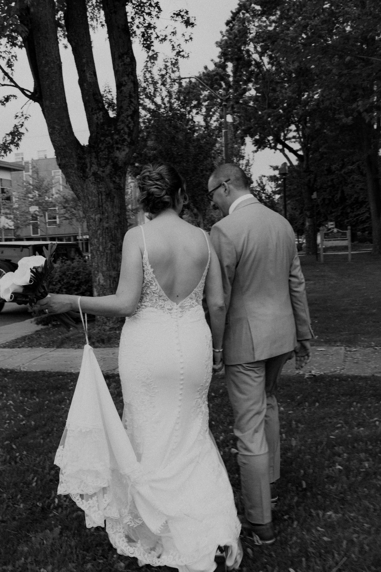 A black and white photo of a bride in a backless lace gown and groom in a tan suit walking under trees.