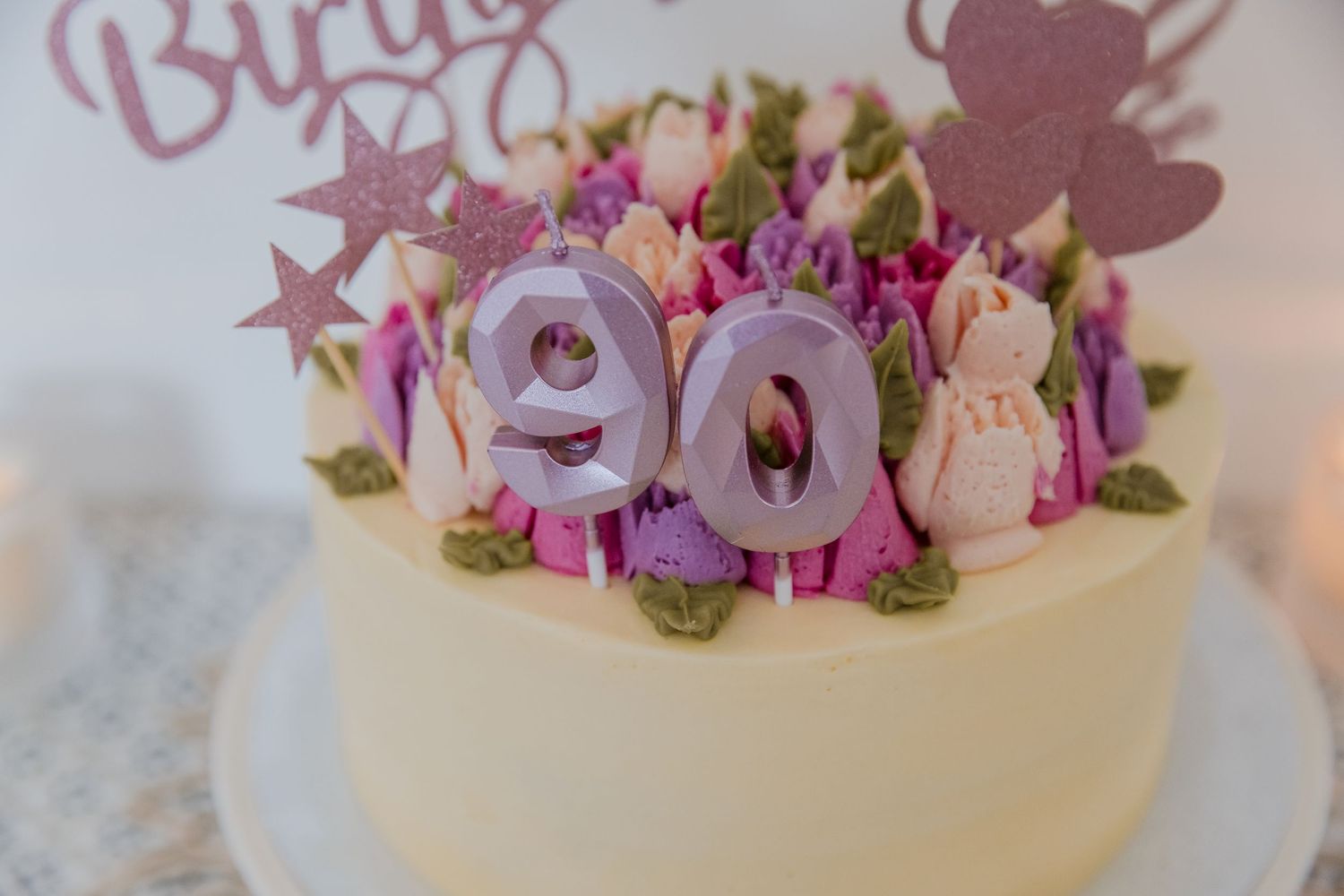 Elegant 90th birthday cake with purple floral decorations, macarons, and glittery number 90 candles on white frosting.