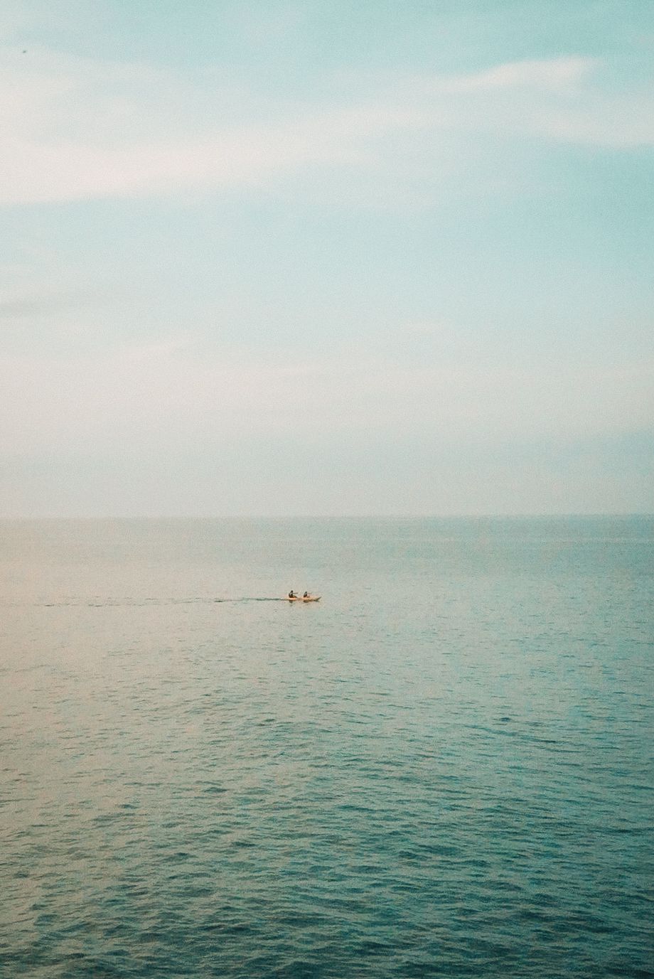 A small boat with two people drifts across a vast, calm turquoise ocean under a soft, hazy pastel sky.