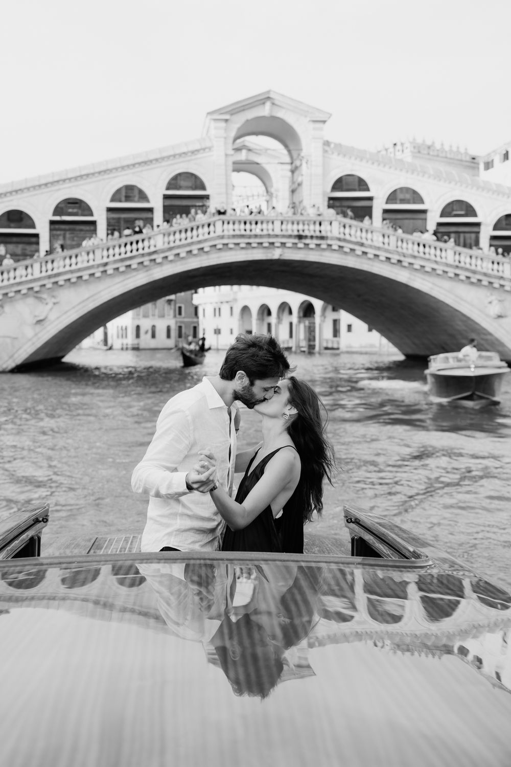 Black and white photo of a couple kissing on a boat in front of the Rialto Bridge in Venice, Italy.