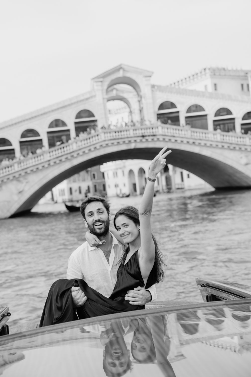 Black and white photo of a smiling couple on a boat in Venice with the Rialto Bridge in the background.