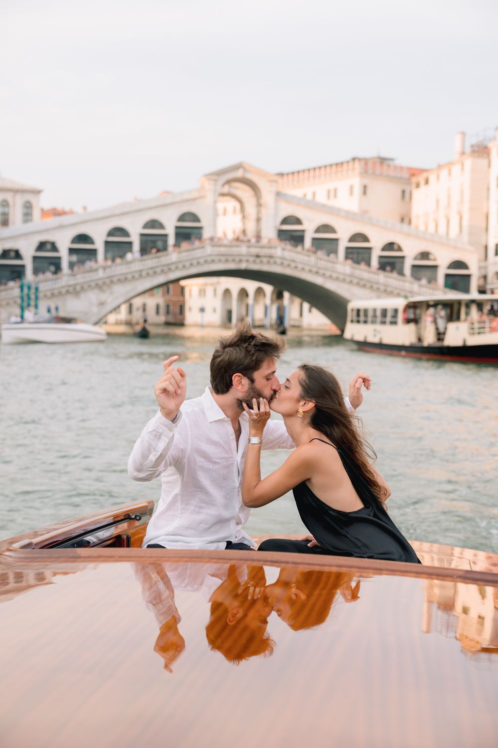 Romantic couple kissing on a boat in Venice's Grand Canal with the iconic Rialto Bridge in the background.