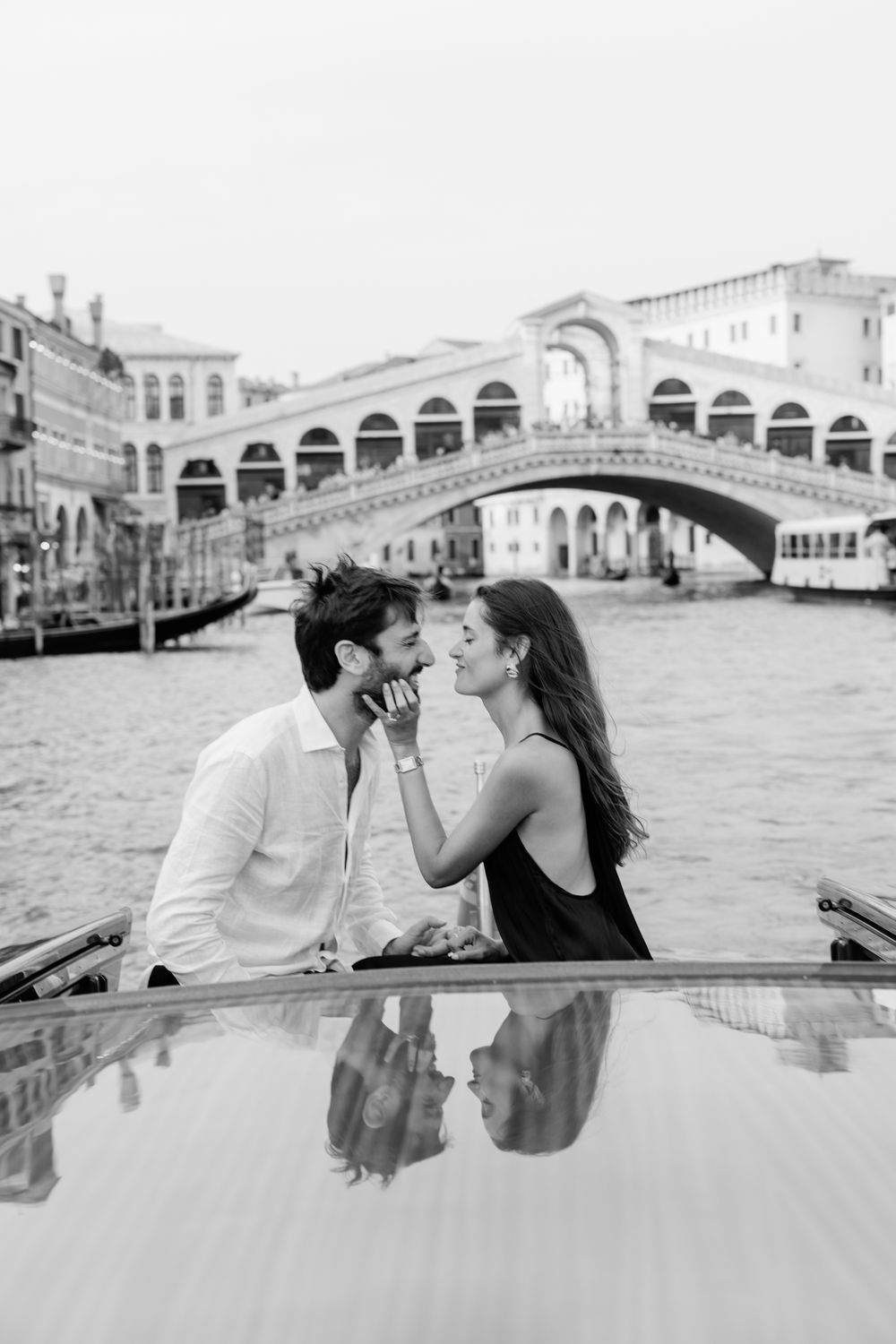 Black and white photo of a romantic couple on a boat in Venice with the Rialto Bridge in the background.