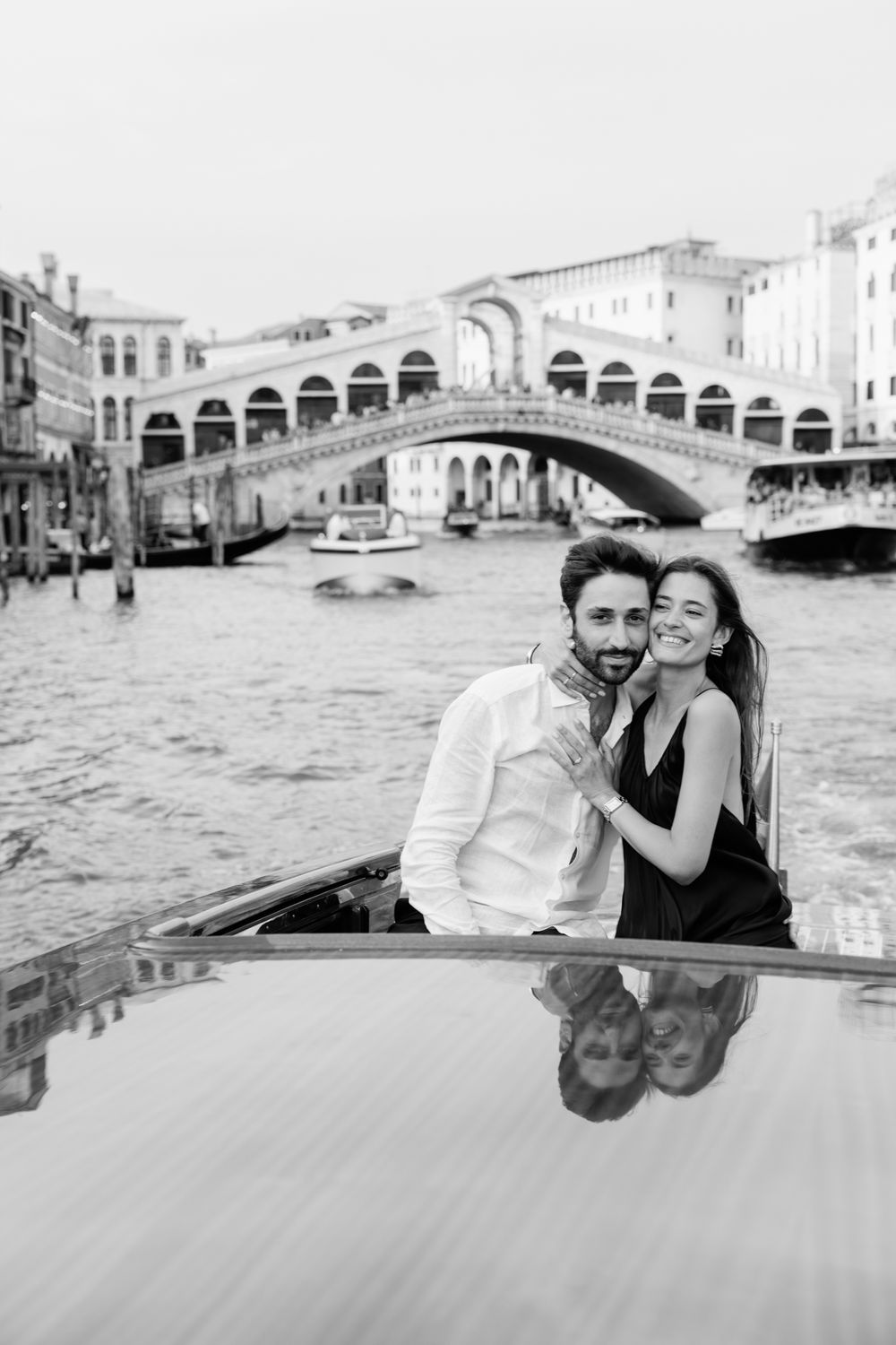Black and white photo of a couple embracing on a gondola with the Rialto Bridge in Venice, Italy in the background.