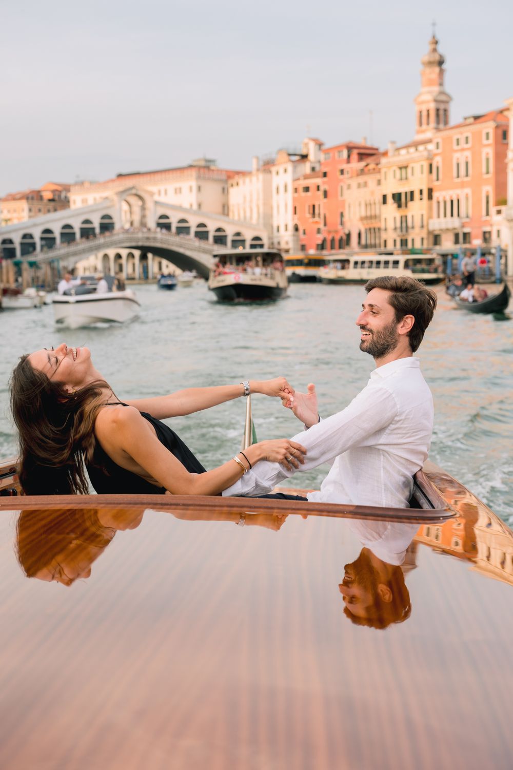 Couple laughing and holding hands on a wooden boat on the Grand Canal near Rialto Bridge in Venice, Italy.