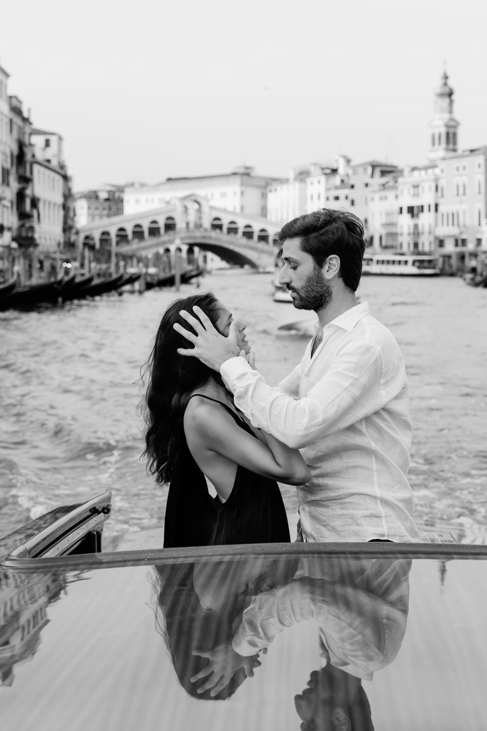 Black and white photo of a romantic couple embracing on a boat in Venice with the Rialto Bridge in the background.