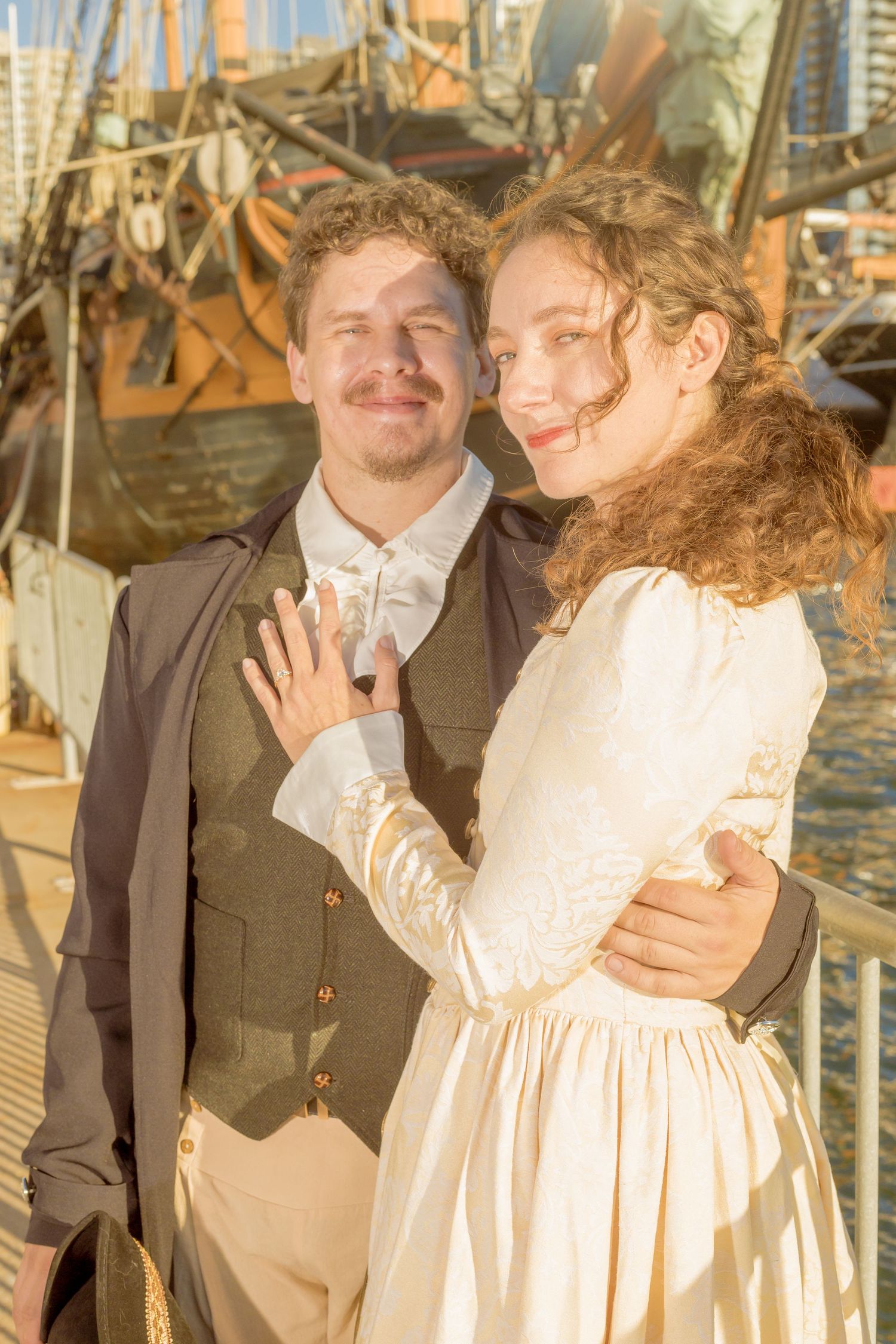 Couple in Victorian period costumes embracing near a historic ship at golden hour.