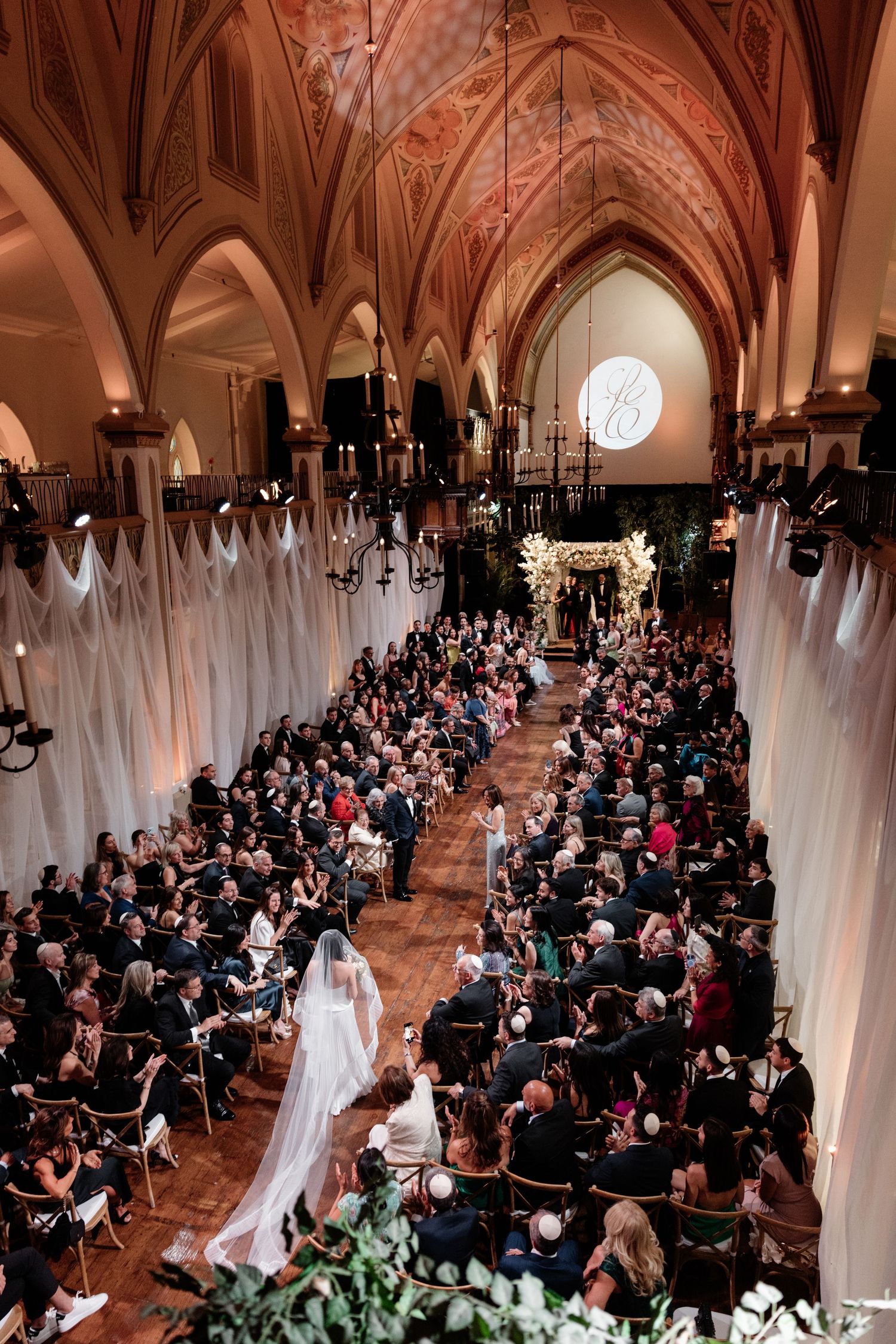 Bride walks down the aisle of an ornate gothic church filled with wedding guests, white draping, and a glowing monogram.