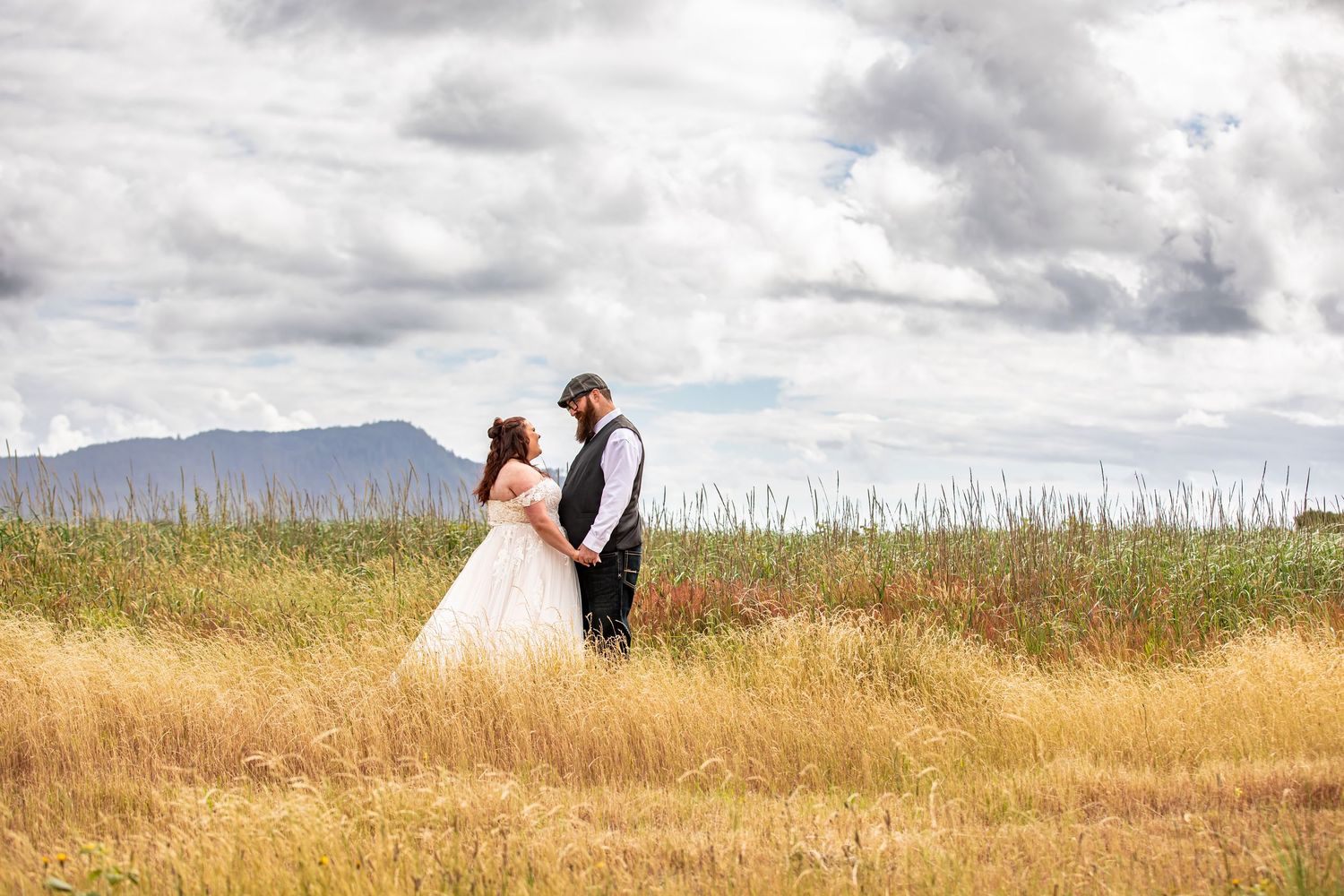 Bride in lace ballgown holds groom's hand walking through green meadow, smiling back at camera on wedding day.