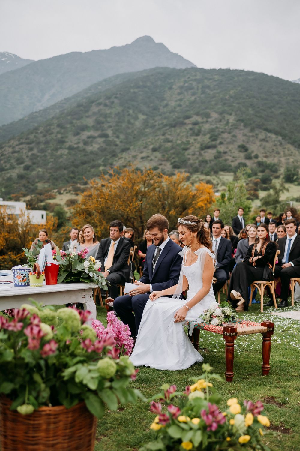 Bride and groom seated during outdoor wedding ceremony with mountain backdrop and colorful flowers.