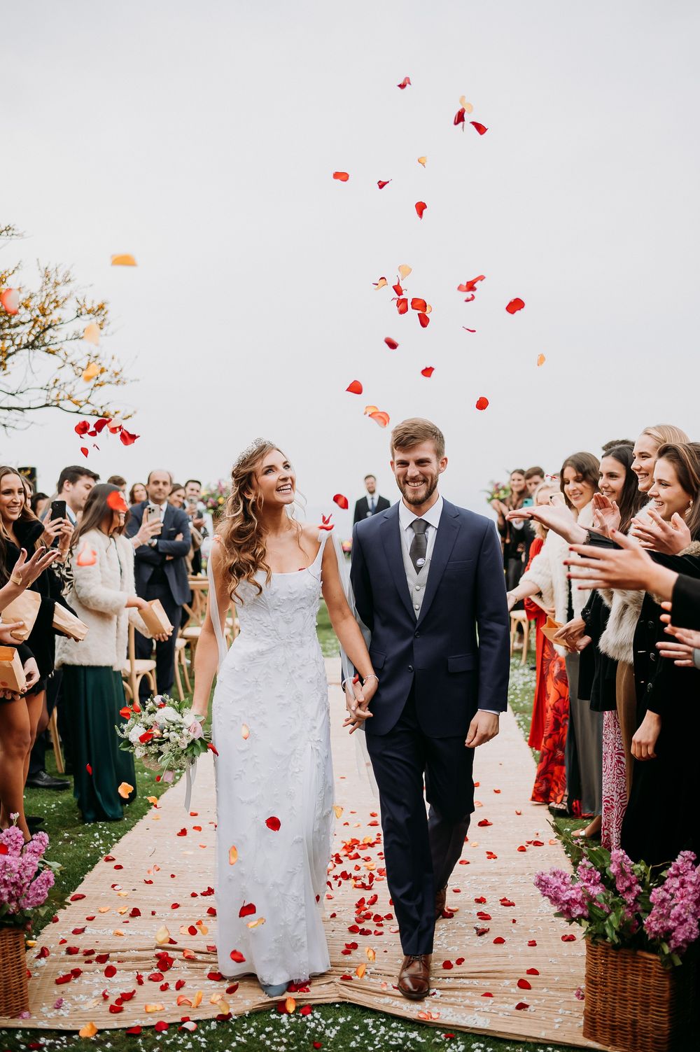 Newlyweds walk down the aisle as guests toss red rose petals at their outdoor wedding ceremony.