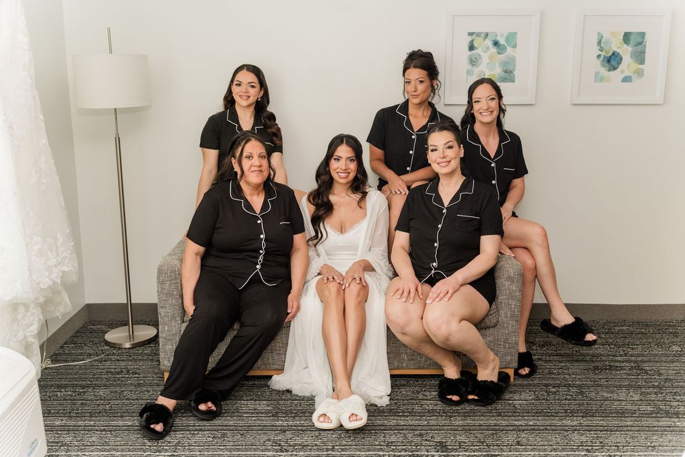 Bride in white robe poses with five bridesmaids in black pajamas, smiling together in a bright hotel room.