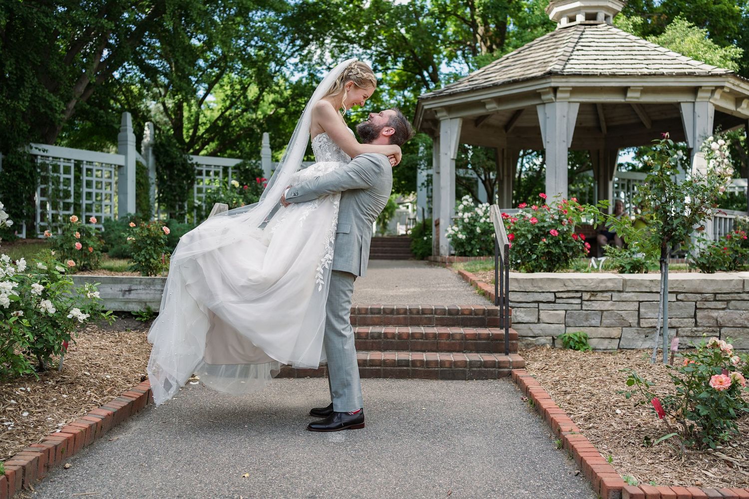 Groom lifting bride near a gazebo surrounded by roses in a beautiful garden setting.