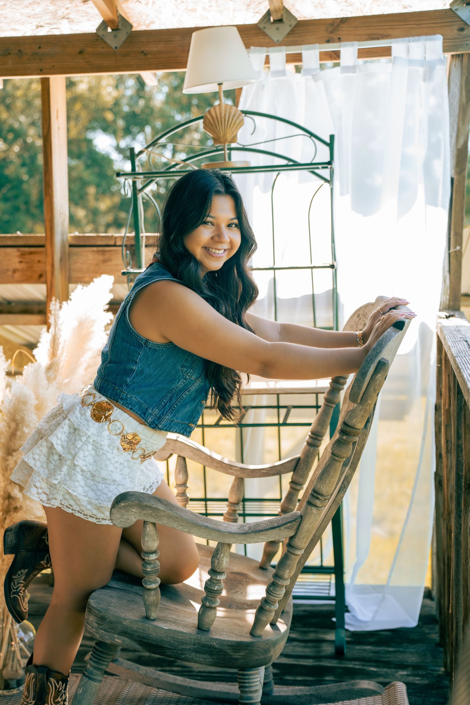 Young woman in denim top and white skirt poses on rustic rocking chair in a warmly lit wooden porch setting.