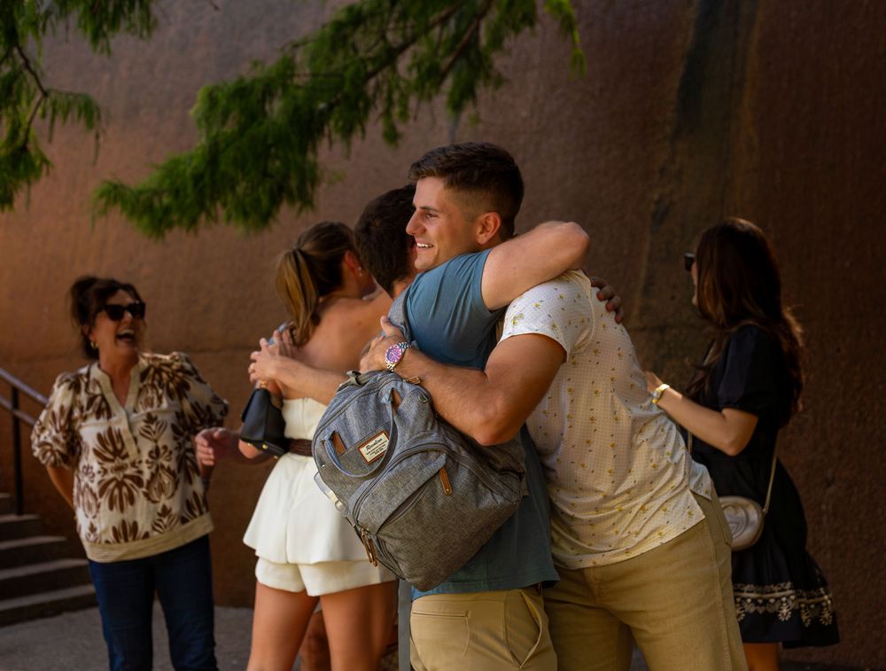 Two young people share a warm embrace outdoors near a brick wall, with others standing nearby in casual summer attire.