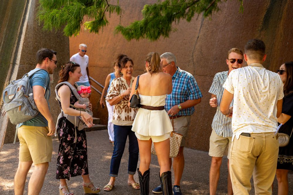 Group of people gathered outdoors near a brick wall in warm weather, socializing and looking at something together.