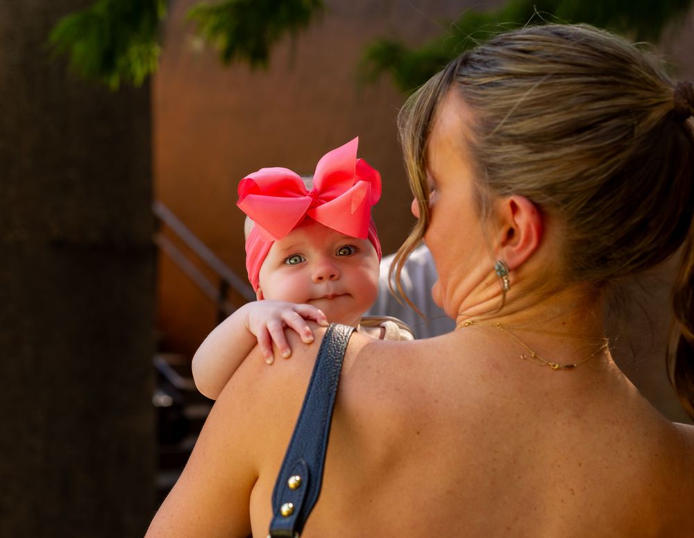 A mother holds her smiling baby girl wearing a large pink bow headband in warm sunlight.