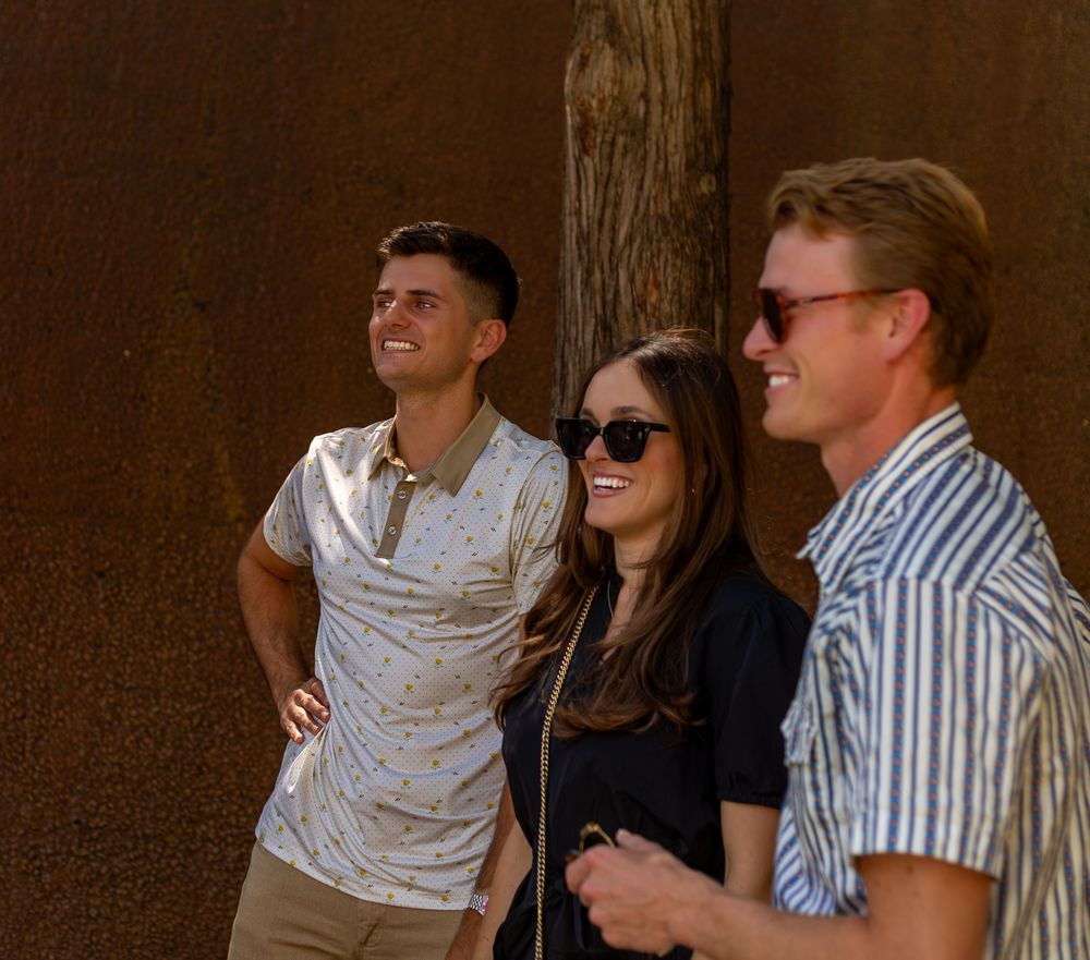 Three smiling young adults standing together outdoors against a rustic brown wall in casual summer attire.
