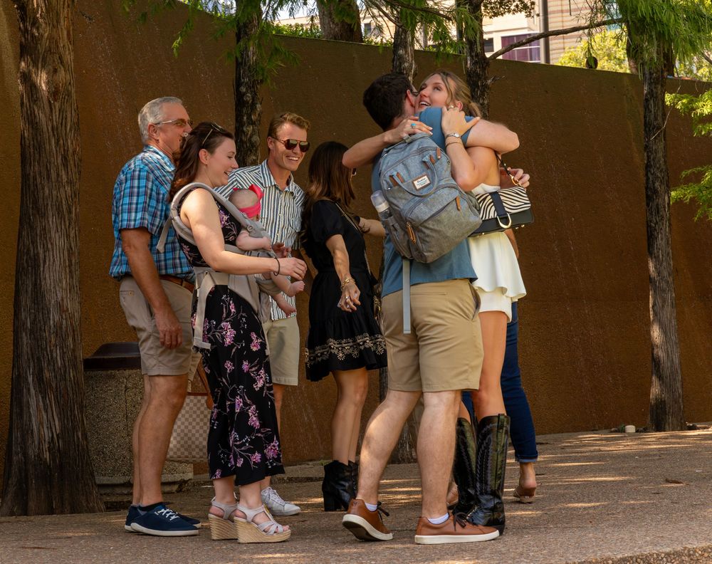Two people embracing warmly while a group of smiling adults watches nearby in an outdoor setting.