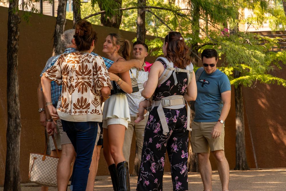 Group of people socializing outdoors near trees, with a woman in a floral dress hugging another.