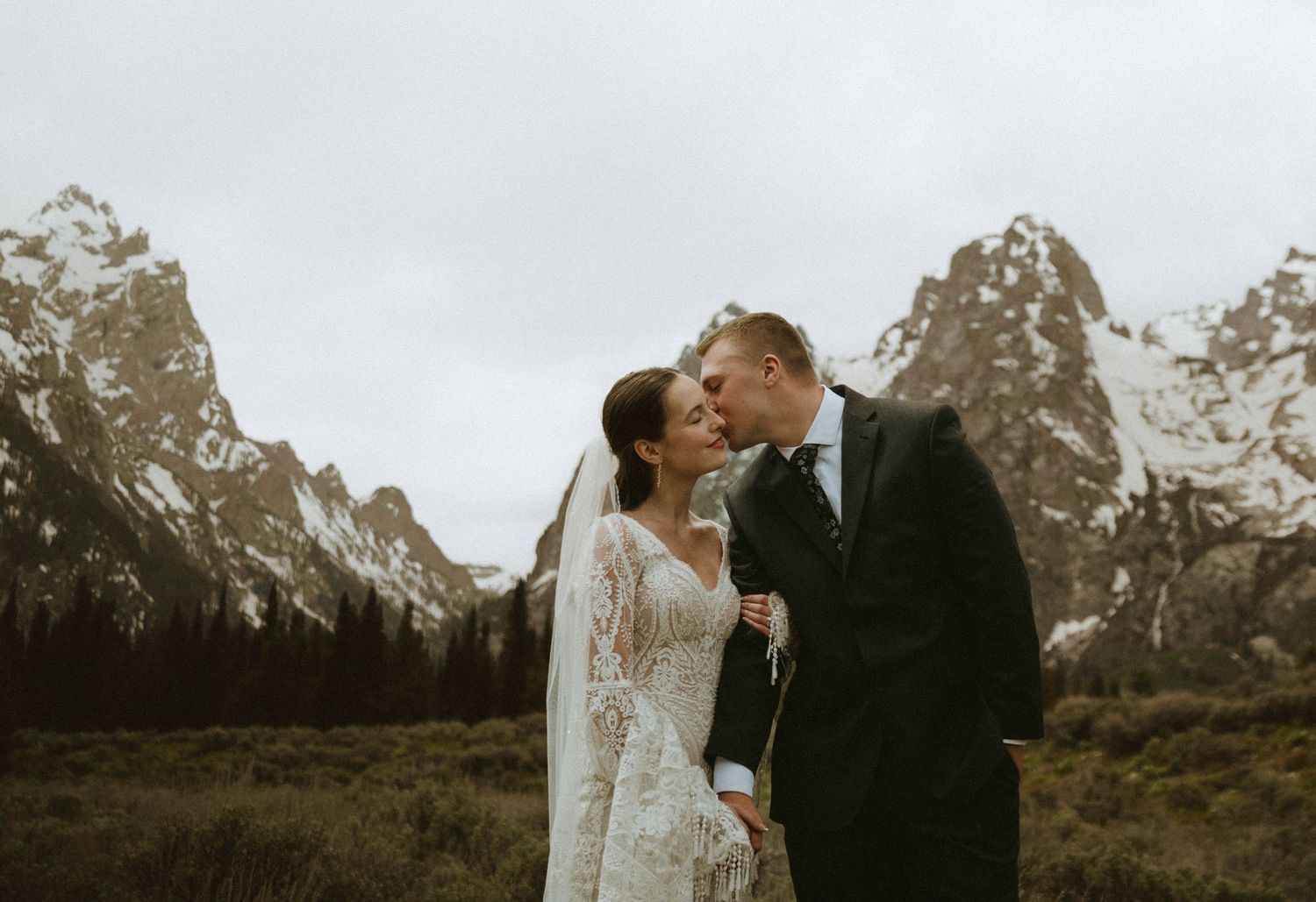 A bride and groom share a romantic kiss in front of snow-capped mountain peaks on their wedding day.